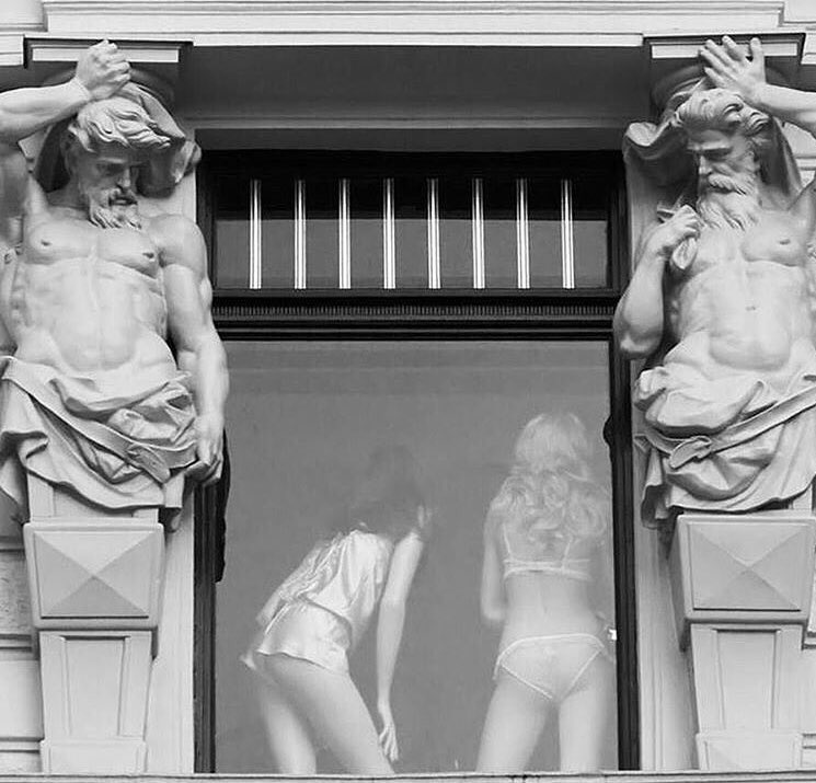 Dos estatuas de Atlantes mirando hacia una ventana en la que se ve a dos mujeres de espaldas en ropa interior
