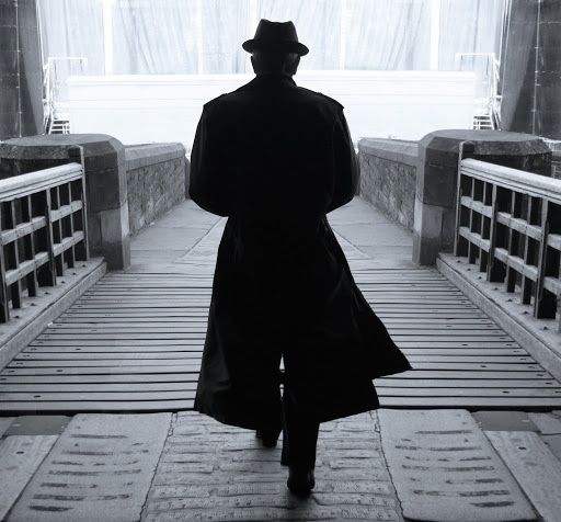Leonard Cohen despaldas con su característico sombrero y una gabardina cruzando un puente de madera. 