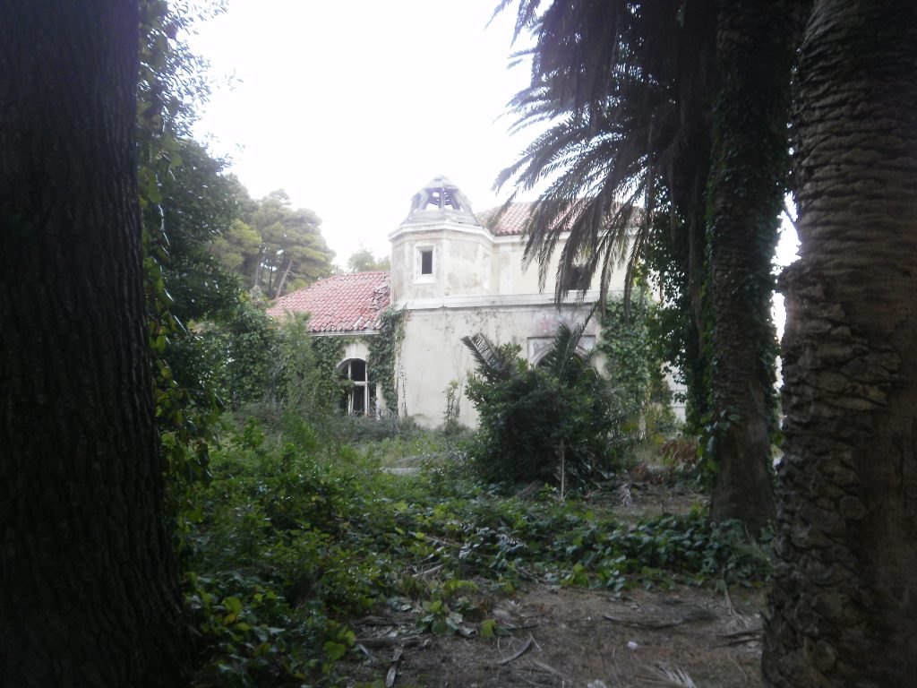 Imagen de un edifico antiguo medio derruido y comido por la maleza que se vislumbra entre unas palmeras en primer plano. (Fotografía de Ignacio J. Dufour García en Kupari (Croacia) en 2012)